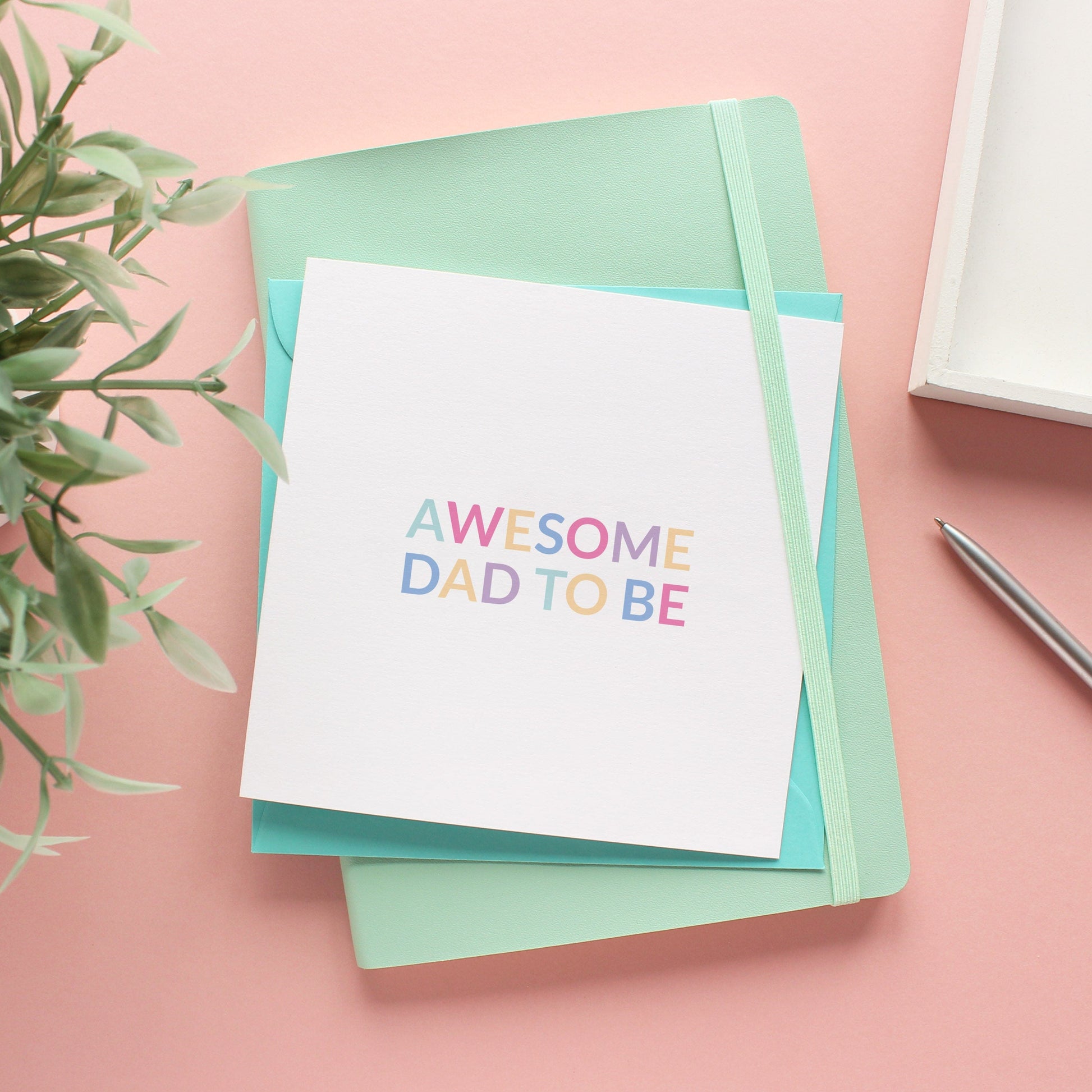 A white greeting card with pastel coloured text that reads awesome dad to be with a turquoise envelope